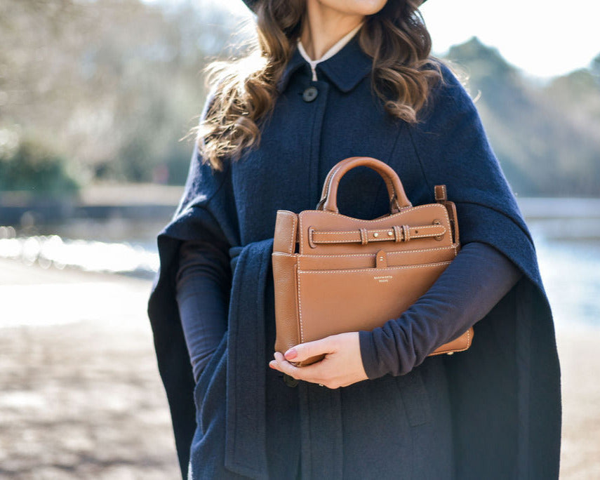Tan leather handbag being held by lady in navy cape infront of a country lake