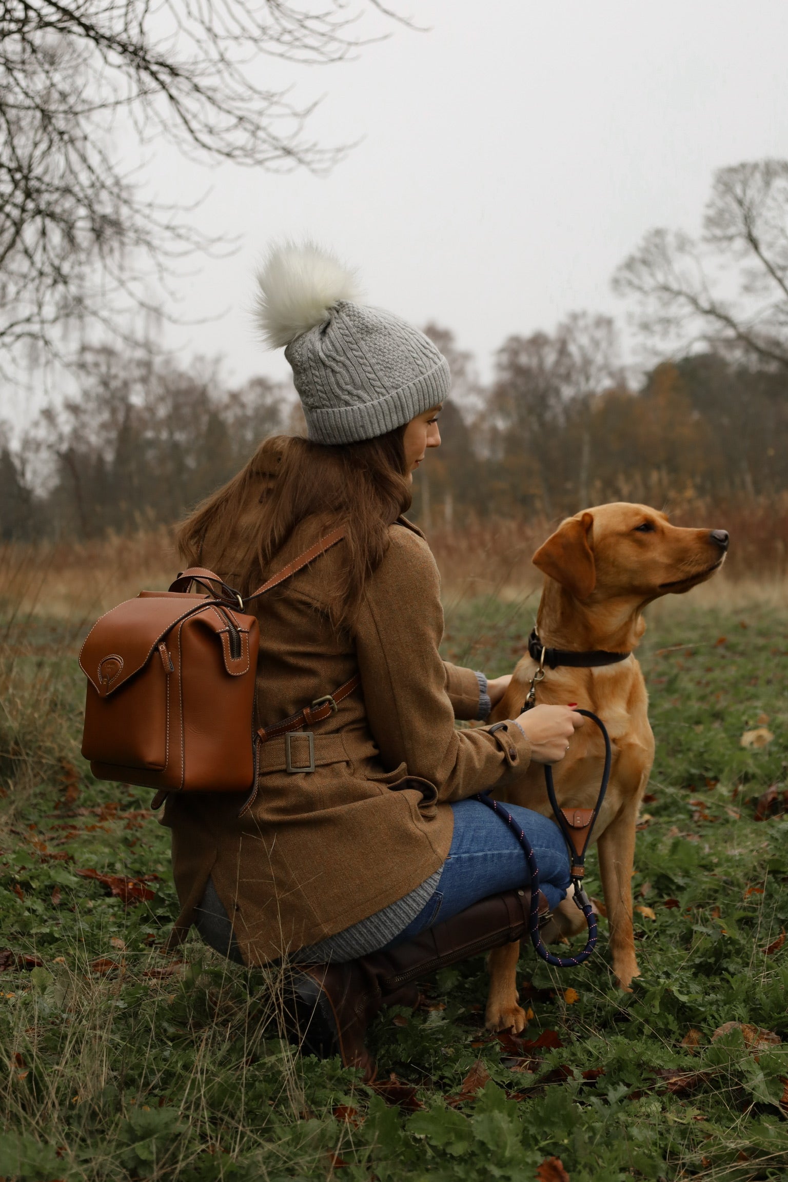 Woman kneeling in a countryside field wearing a tweed coat, grey bobble hat and jeans, carrying a Barkworth Reeve tan leather backpack while holding the lead of a golden labrador dog on an autumn dog walk.
