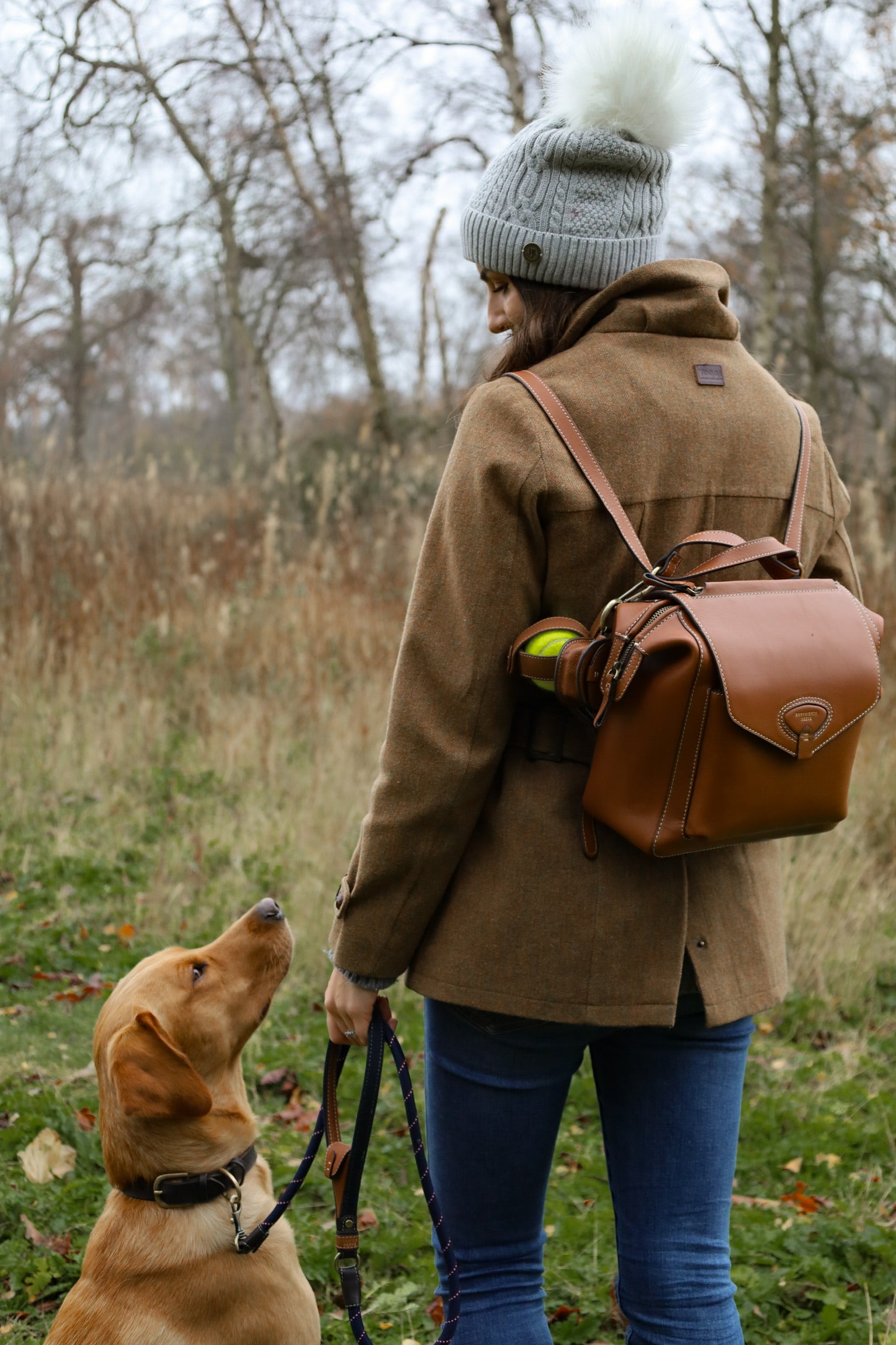 Woman in a brown tweed country coat, jeans and grey Barkworth Reeve bobble hat walking through a field with a Barkworth Reeve tan leather backpack with a tan leather tennis ball holder, while her golden labrador sits at her side on a lead.