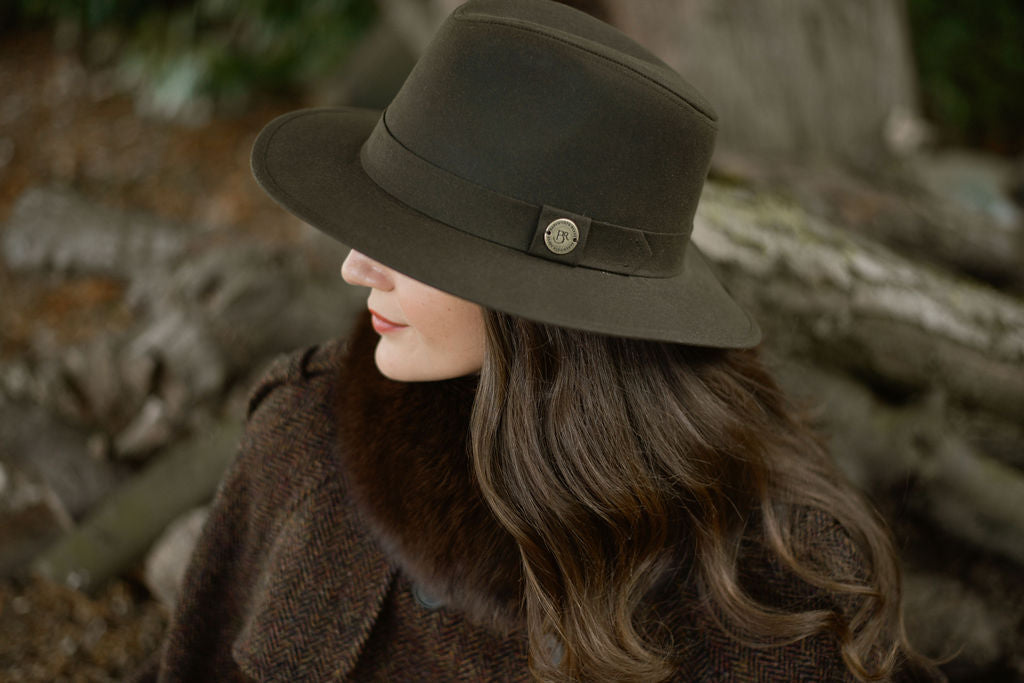 Woman wearing a dark brown fedora hat with a decorative button, standing against a natural background.
