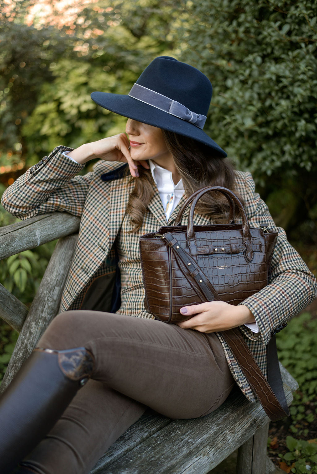 Woman in tweed equestrian attire with a brown leather handbag and navy blue woollen fedora hat sitting on a wooden bench outdoors