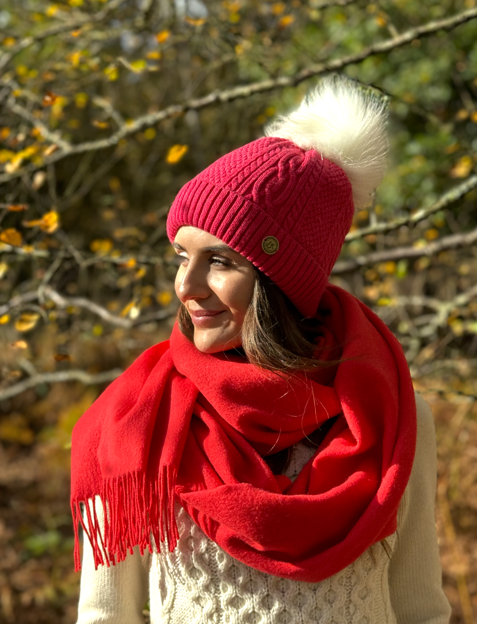 lady wearing raspberry pink bobble hat with white faux fur pom pom and a red scarf , in a autumn wood.