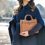 Tan leather handbag being held by lady in navy cape infront of a country lake