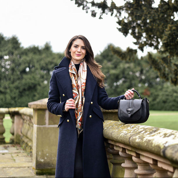 Woman in a navy coat holding a black barkworth reeve saddle bag  in an outdoor setting with greenery.
