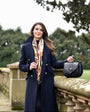 Woman in a navy coat holding a black barkworth reeve saddle bag  in an outdoor setting with greenery.