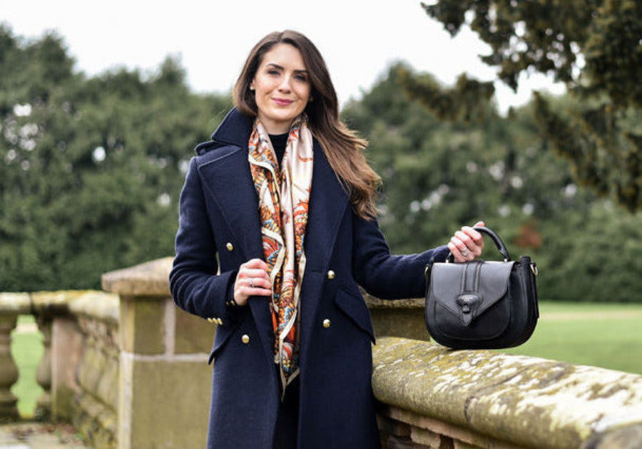 Woman in a navy coat holding a black barkworth reeve saddle bag  in an outdoor setting with greenery.