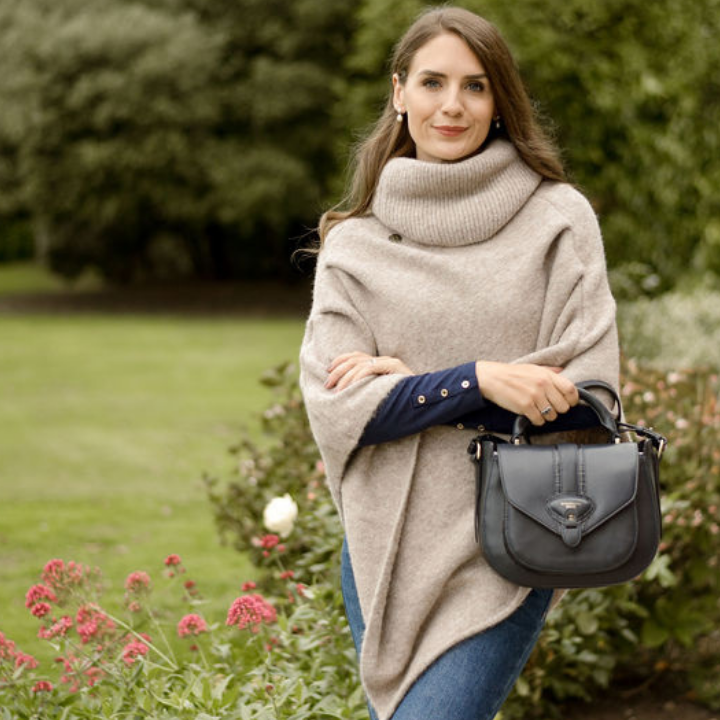 Woman in a beige poncho holding a gray handbag in a garden setting