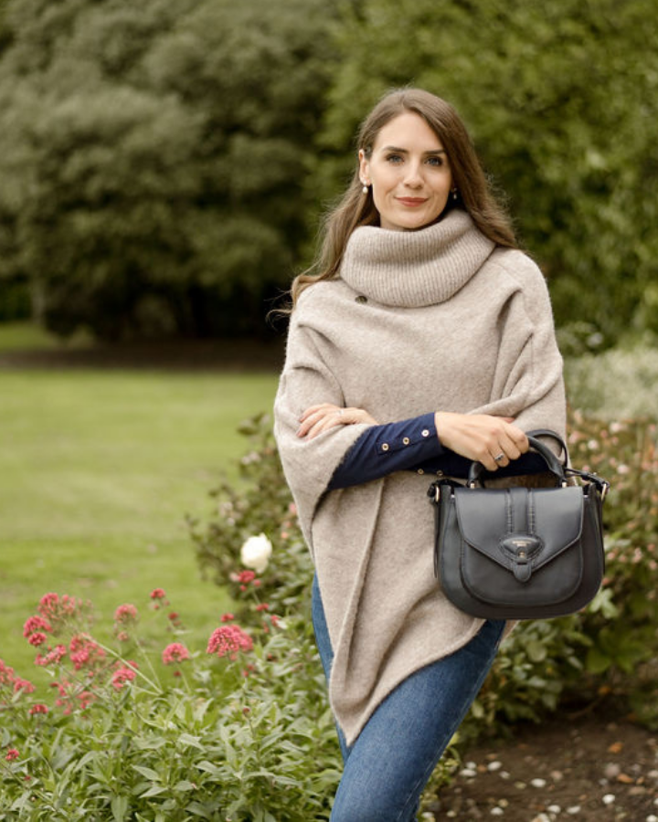 Woman in a beige poncho holding a gray handbag in a garden setting