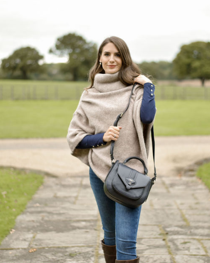 Woman wearing a beige poncho and blue jeans, holding a gray handbag in an outdoor setting.