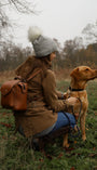Woman kneeling in a countryside field wearing a tweed coat, grey bobble hat and jeans, carrying a Barkworth Reeve tan leather backpack while holding the lead of a golden labrador dog on an autumn dog walk.