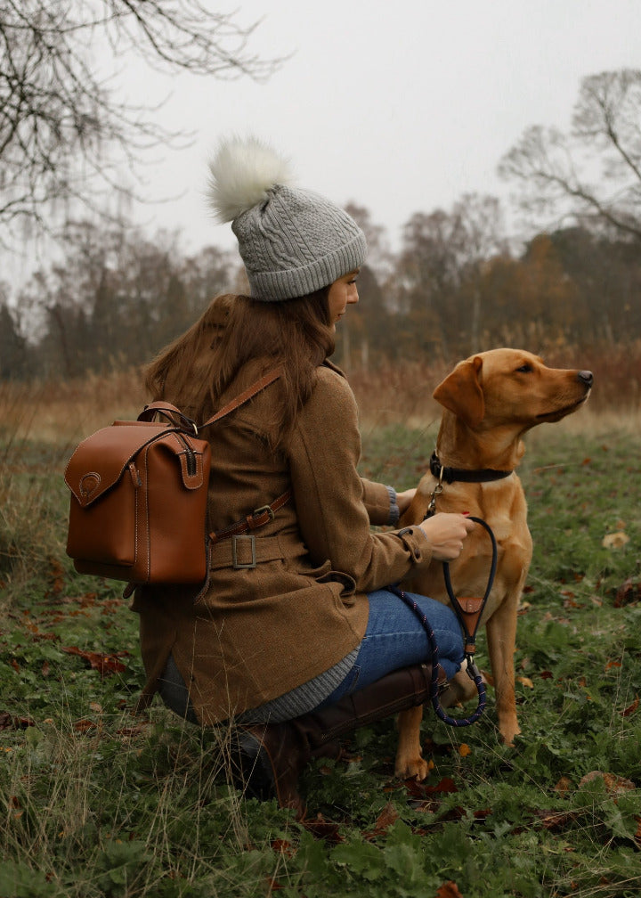 Woman kneeling in a countryside field wearing a tweed coat, grey bobble hat and jeans, carrying a Barkworth Reeve tan leather backpack while holding the lead of a golden labrador dog on an autumn dog walk.