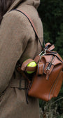 Close-up of a woman in a brown tweed country coat wearing a Barkworth Reeve tan leather  walking backpack with contrast stitching, showing a tennis dog ball holder in a side strap and zipped top pocket detail.