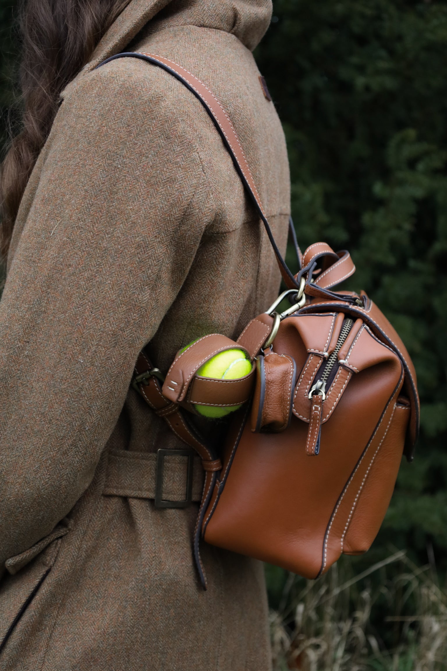 Close-up of a woman in a brown tweed country coat wearing a Barkworth Reeve tan leather  walking backpack with contrast stitching, showing a tennis dog ball holder in a side strap and zipped top pocket detail.