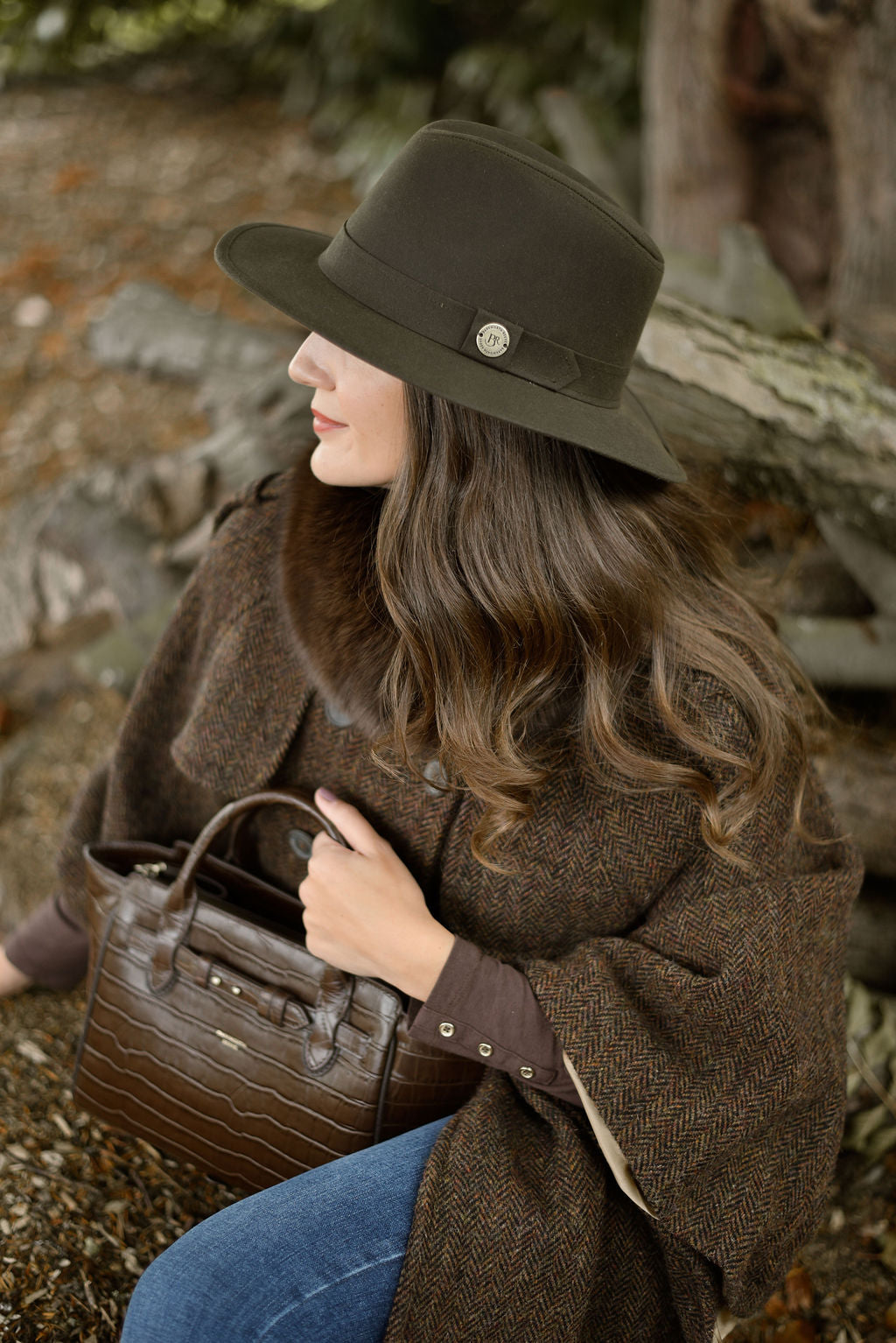 Woman in a brown coat and hat holding a brown handbag, sitting on a log.