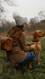 WOMAN AND DOG IN WOODLAND. WOMAN KNEELING WEARING GREY BOBBLE HAT AND LEATHER RUCKSACK