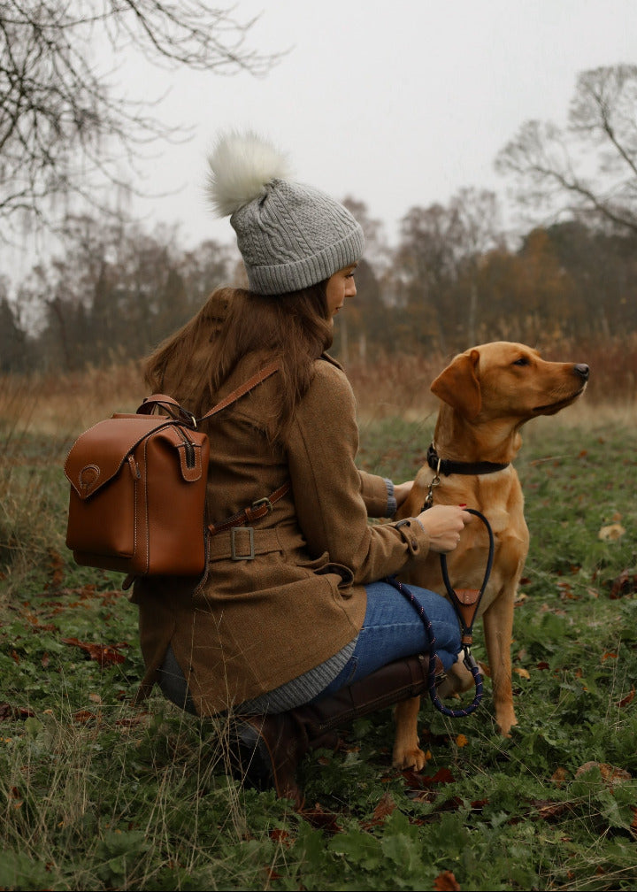 WOMAN AND DOG IN WOODLAND. WOMAN KNEELING WEARING GREY BOBBLE HAT AND LEATHER RUCKSACK