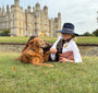 Woman and golden retriver dog sitting on grass in front of Burghely House.  Woman is wearing country style navy fedora hat and tan croc leather clutch bag