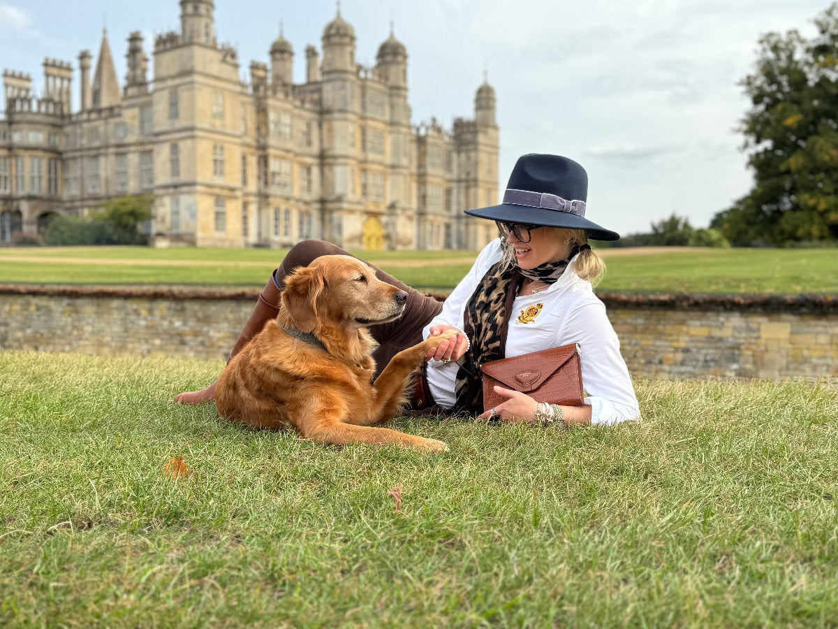 Woman and golden retriver dog sitting on grass in front of Burghely House.  Woman is wearing country style navy fedora hat and tan croc leather clutch bag