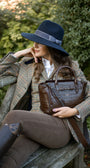 Woman in tweed equestrian attire with a brown leather handbag and navy blue woollen fedora hat  sitting on a wooden bench outdoors