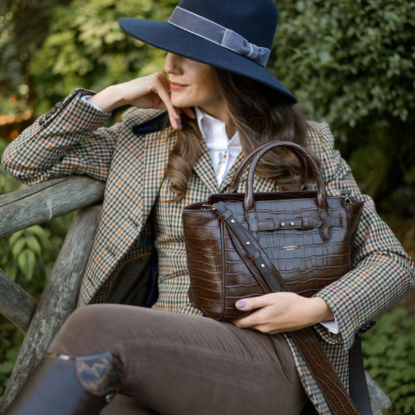 Woman in tweed equestrian attire with a brown leather handbag and navy blue woollen fedora hat  sitting on a wooden bench outdoors