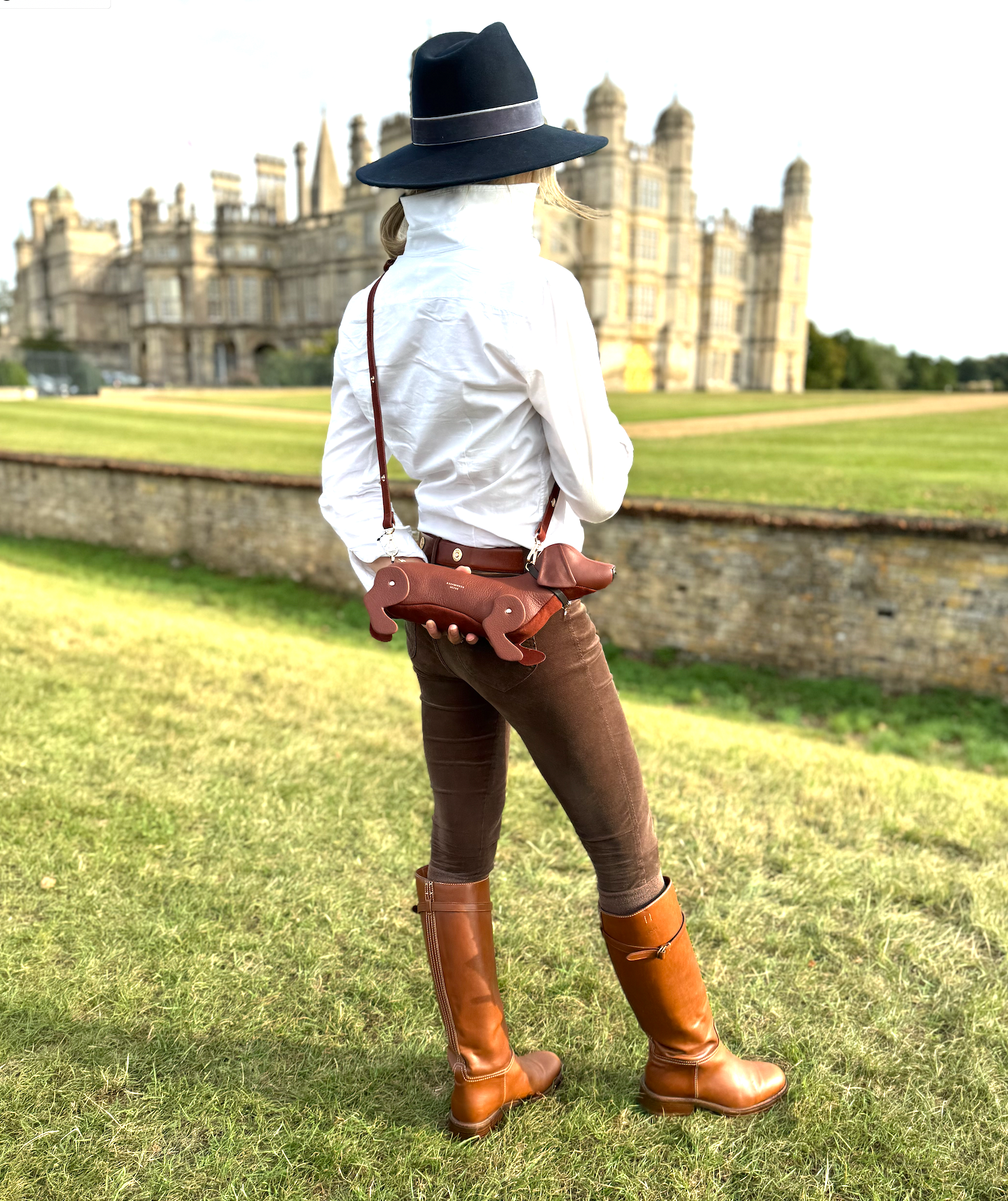 lady in formal country attire with a hat and boots and sausage dog bagstanding in front of a large castle.