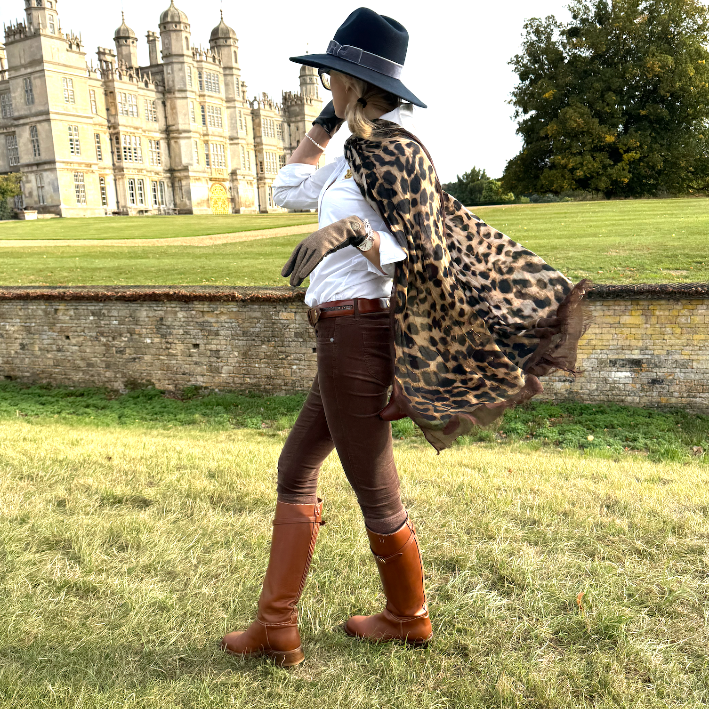woman in a leopard print scarf, brown boots and a navy hat standing in a grassy field with a stately home in the background.