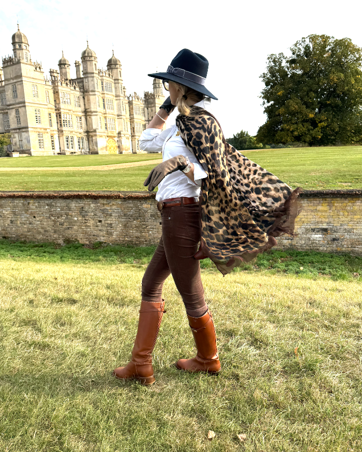 woman in a leopard print scarf, brown boots and a navy hat standing in a grassy field with a stately home in the background.