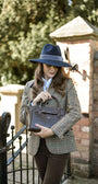 Woman in tweed check equestrian style jacket and navy blue fedora hat holding a chocolate croc leather handbag, standing in front of a gate.