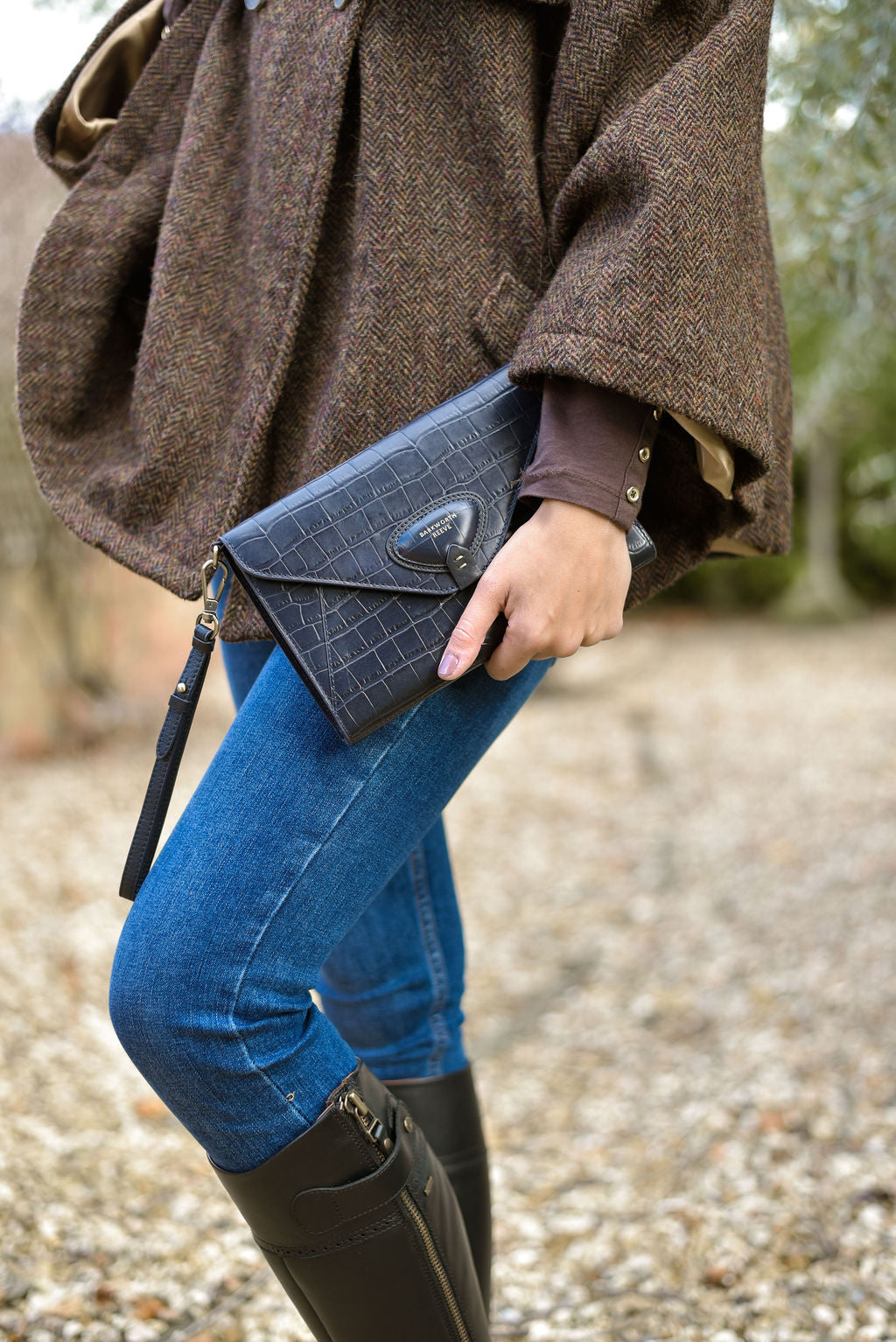 woman wearing jeans and tweed, holding a navy blue croc skin leather clutch bag with a blurred natural background