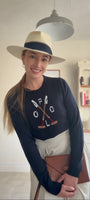 Woman wearing a navy sweater with polo design, white skirt, and wide-brimmed hat indoors.
