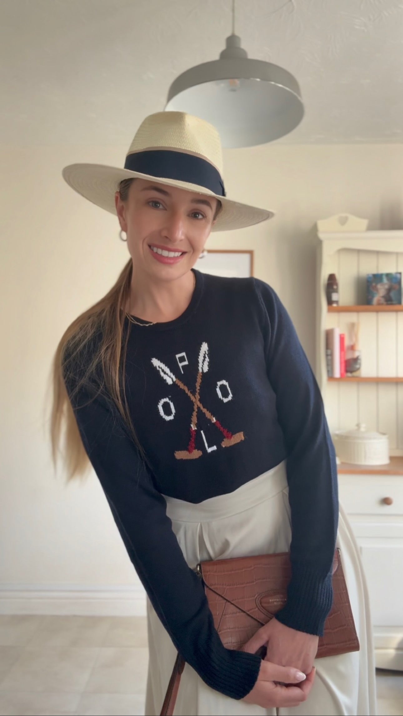 Woman wearing a navy sweater with polo design, white skirt, and wide-brimmed hat indoors.