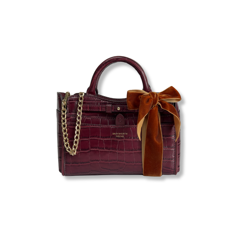 A burgundy crocodile-embossed leather handbag with a top handle, gold chain strap, and a large rust-brown velvet bow on the front, shown on a plain white background.