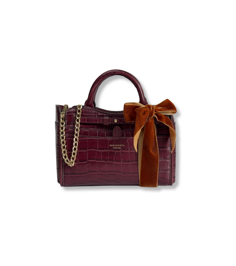 A burgundy crocodile-embossed leather handbag with a top handle, gold chain strap, and a large rust-brown velvet bow on the front, shown on a plain white background.