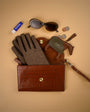 Tan Brown leather clutch bag open with tweed gloves, sunglasses, and other items on a beige background