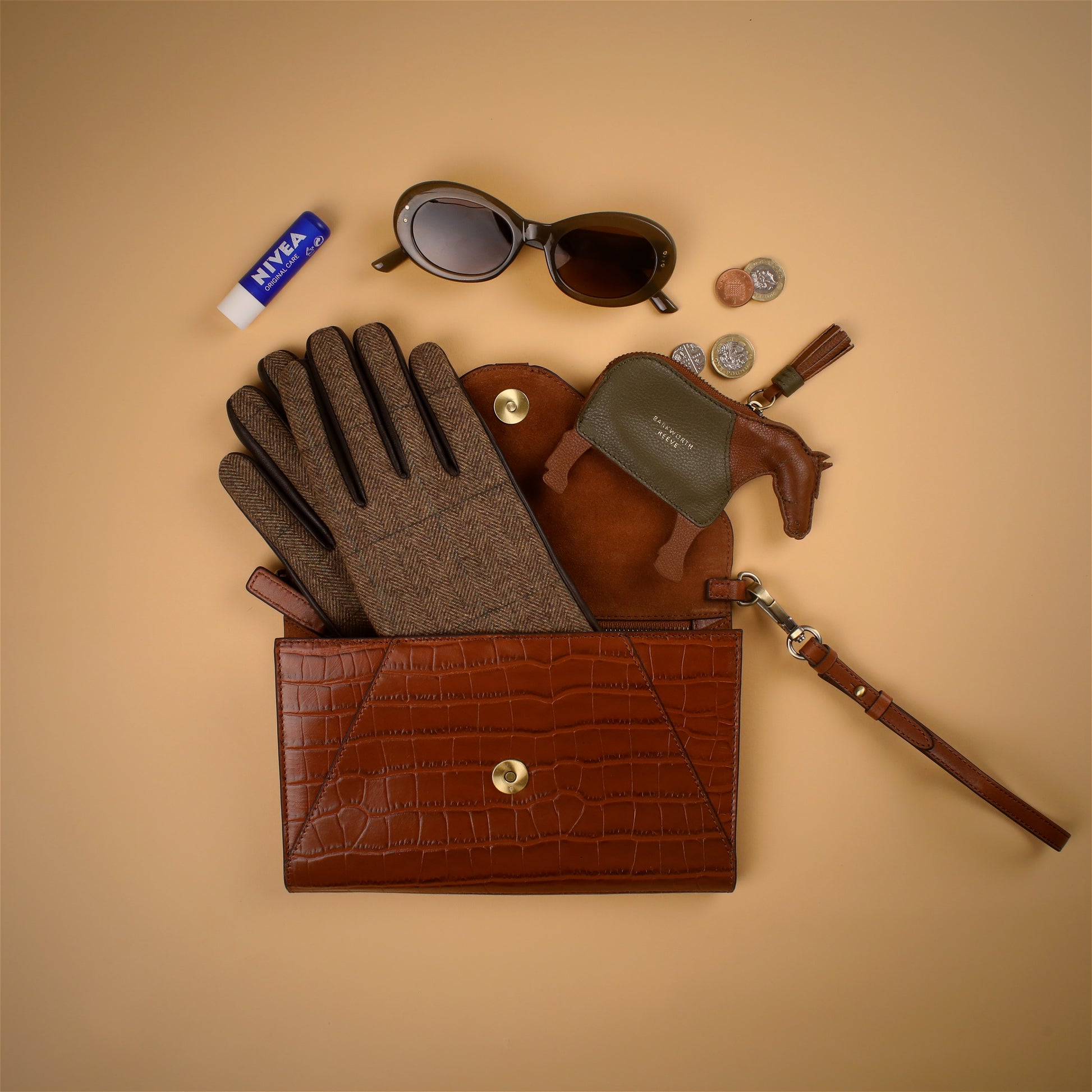 Tan Brown leather clutch bag open with tweed gloves, sunglasses, and other items on a beige background
