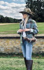 A woman stands outdoors on a grassy lawn wearing a wide-brimmed brown hat with feathers, a green and blue plaid fringed shirt jacket, a white shirt, blue jeans, and tall dark brown riding boots. She is wearing a brown leather crossbody bag with fringe and a buckle strap, set against a background of a stone wall, green fields, and trees under a cloudy sky.