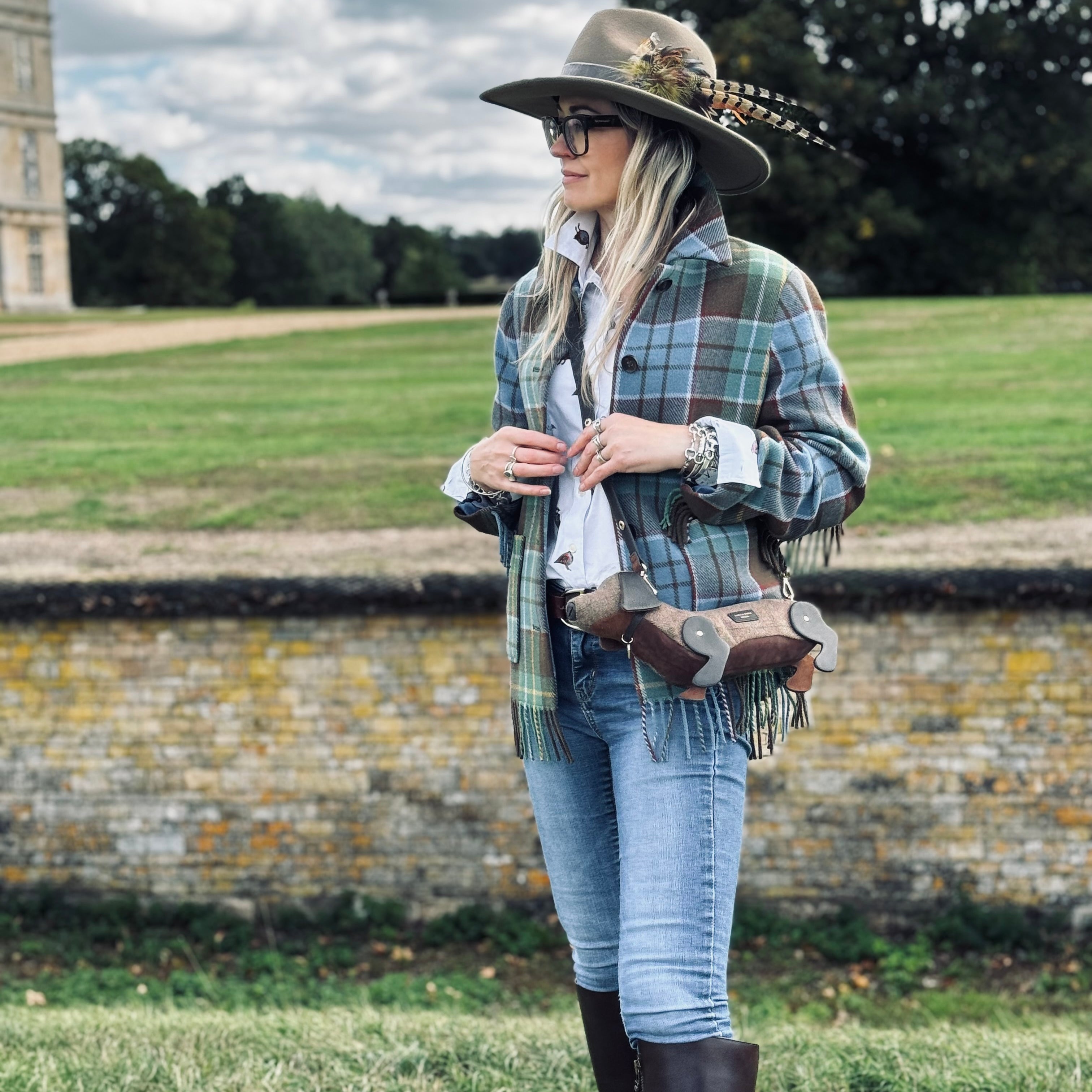 A woman stands outdoors on a grassy lawn wearing a wide-brimmed brown hat with feathers, a green and blue plaid fringed shirt jacket, a white shirt, blue jeans, and tall dark brown riding boots. She is wearing a brown leather crossbody bag with fringe and a buckle strap, set against a background of a stone wall, green fields, and trees under a cloudy sky.