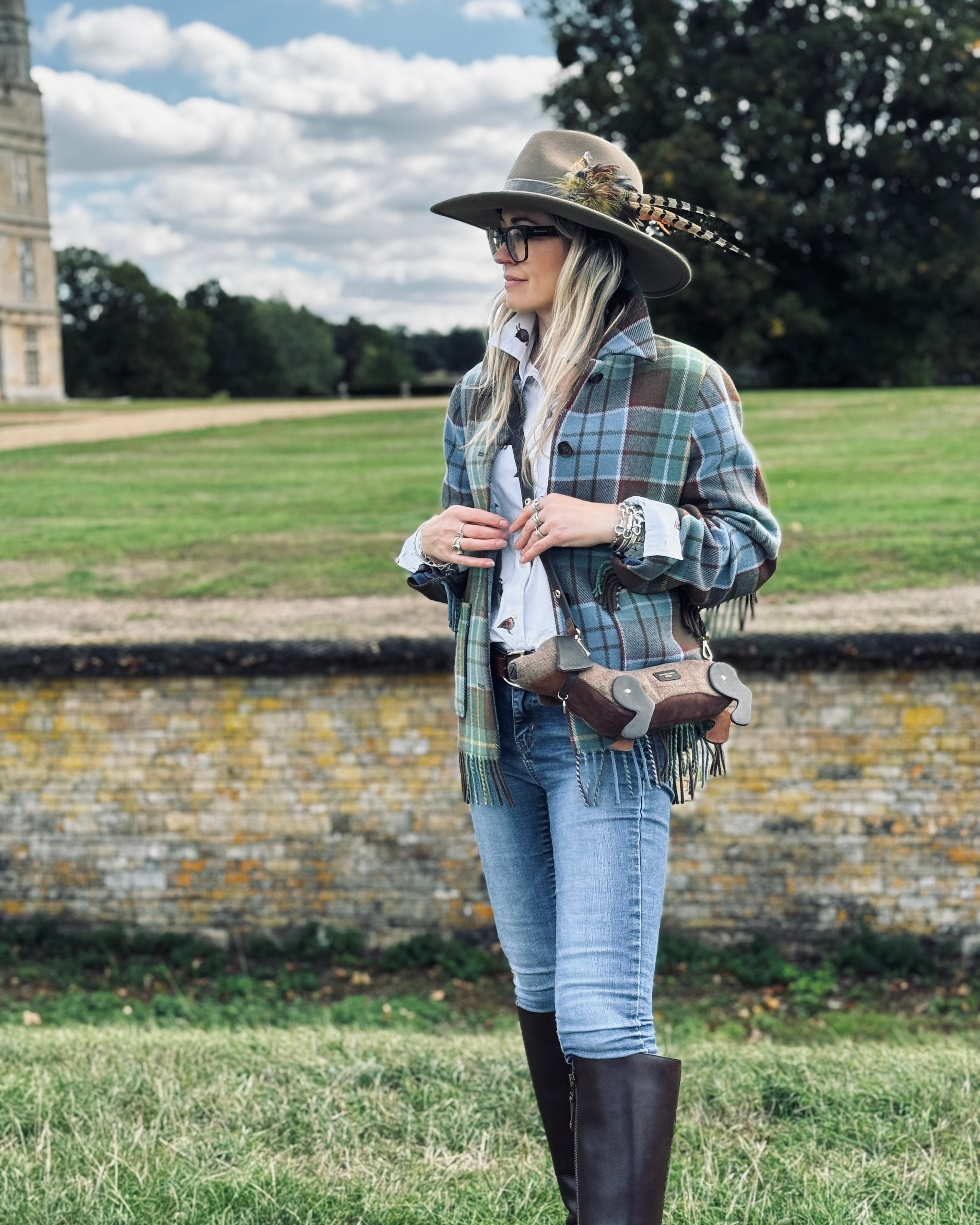 A woman stands outdoors on a grassy lawn wearing a wide-brimmed brown hat with feathers, a green and blue plaid fringed shirt jacket, a white shirt, blue jeans, and tall dark brown riding boots. She is wearing a brown leather crossbody bag with fringe and a buckle strap, set against a background of a stone wall, green fields, and trees under a cloudy sky.