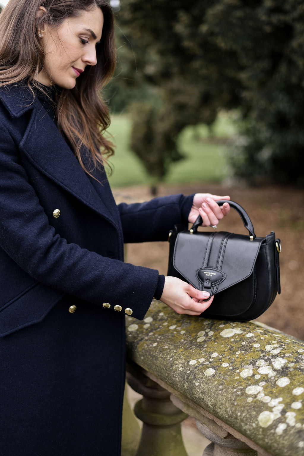 Woman in a navy coat holding a black handbag outdoors.