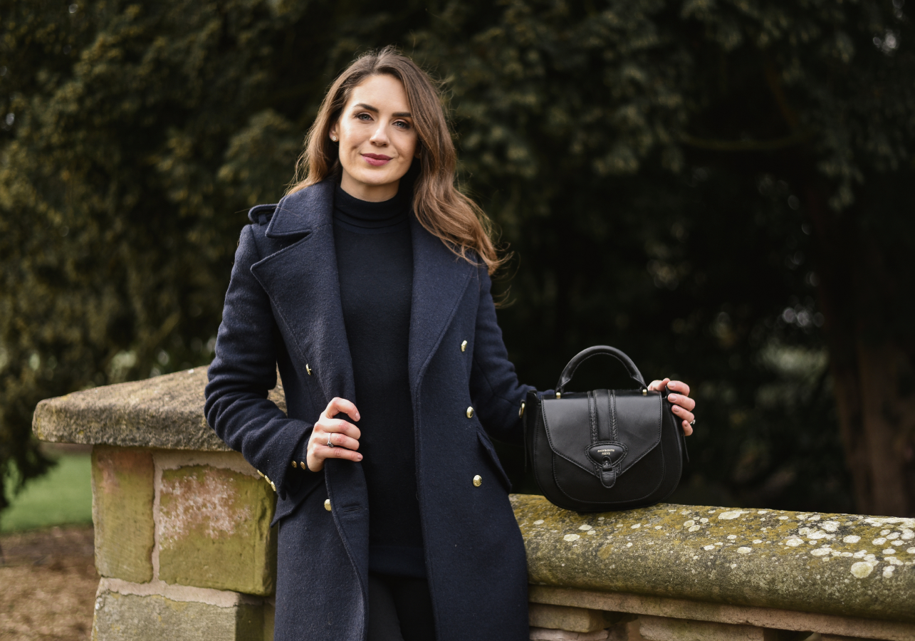 Woman in a dark navy coat holding a black handbag against a blurred natural background