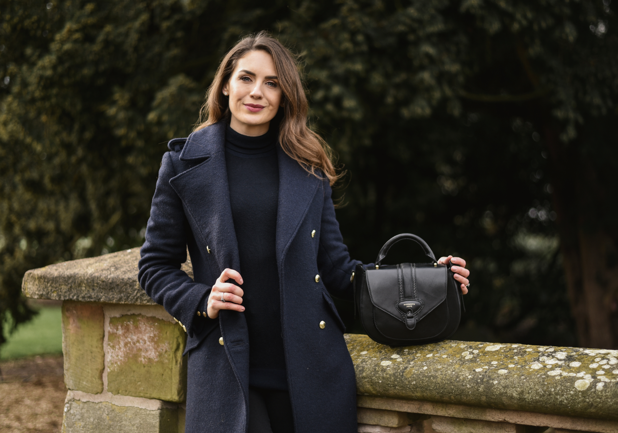 Woman in a dark navy coat holding a black handbag against a blurred natural background