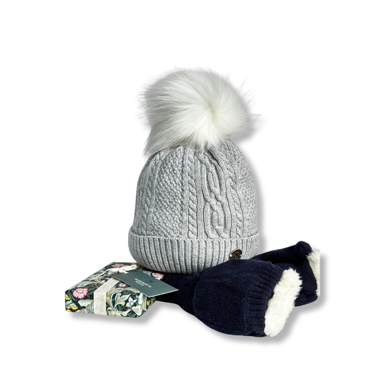 A light gray cable-knit beanie with a large white faux-fur pom-pom, shown with navy blue mittens featuring white trim and a small floral gift box on a white background.