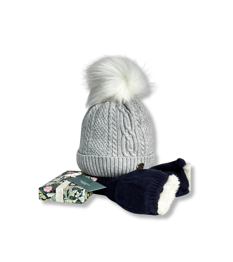 A light gray cable-knit beanie with a large white faux-fur pom-pom, shown with navy blue mittens featuring white trim and a small floral gift box on a white background.