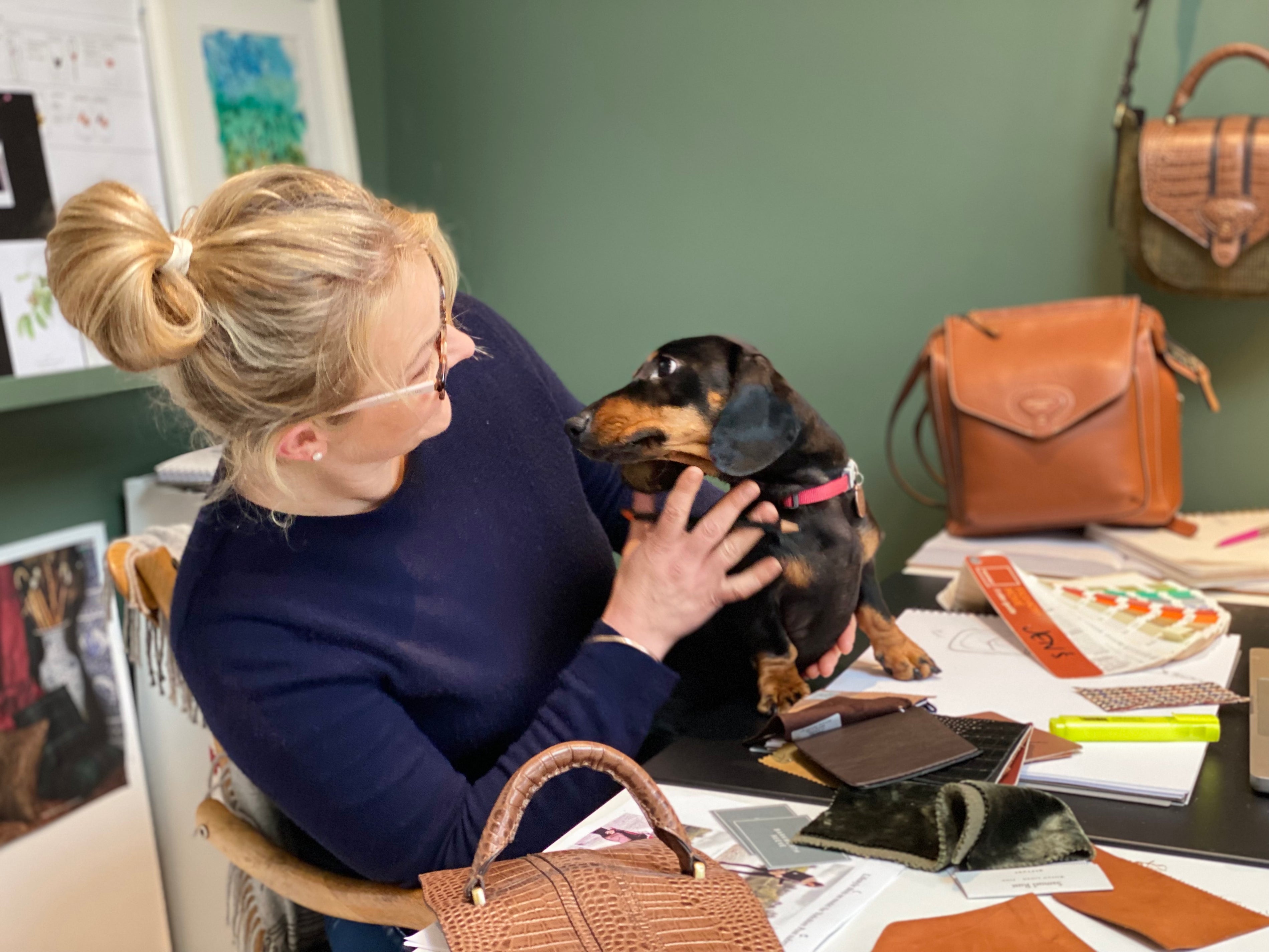 woman and sausage dog at a desk with handbags in background
