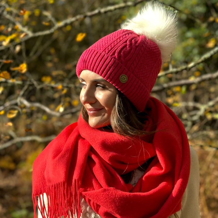 lady wearing raspberry pink bobble hat with white faux fur pom pom and a red scarf , in a autumn wood.
