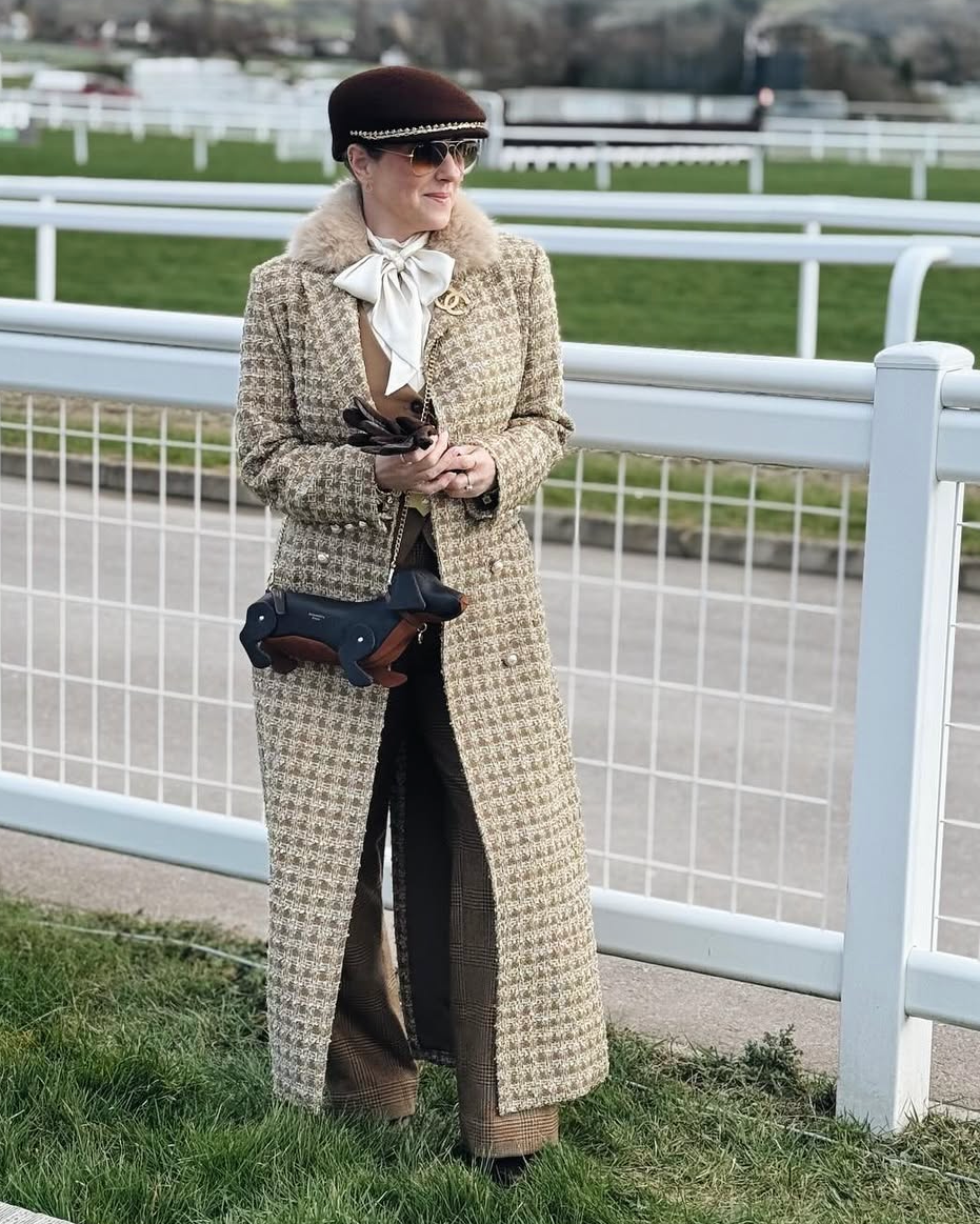 Woman in a long coat standing on a grassy area with a white fence in the background