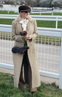 Woman in a long coat standing on a grassy area with a white fence in the background