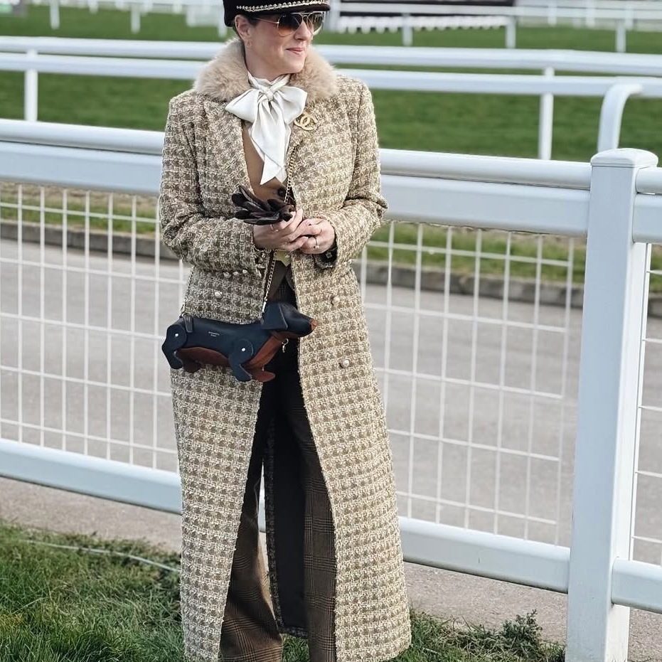 Woman in a long coat standing on a grassy area with a white fence in the background