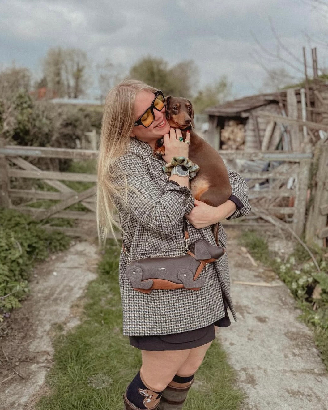 Woman holding a small dog outdoors with a wooden fence and trees in the background